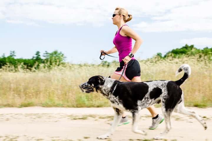Photo of Woman Jogging On Sand With Muzzled Dog Min